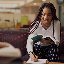 An international student sat in a collaborative area of CCE1; they are laughing, holding a book in one hand and making notes in the other.