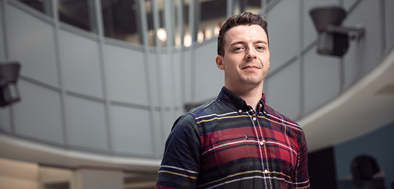Solo shot of a Postgraduate student in a checkered shirt in the CCE1 entry way. He is looking directly at the camera.
