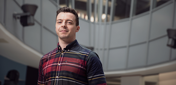 Solo shot of a Postgraduate student in a checkered shirt in the CCE1 entry way. He is looking directly at the camera.
