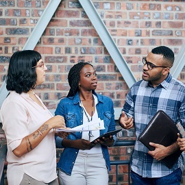 Three colleagues stood in office setting whilst holding folders and talking 