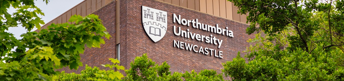 A silver sign that reads 'Northumbria University Northumbria' on a wall, leaves in the forefront 