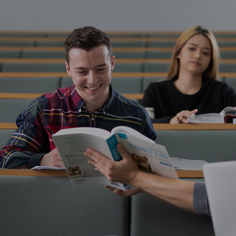 A student is passing a textbook to another student who is taking notes and smiling.