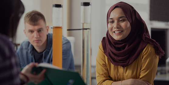 Three students in a lab. One is holding a textbook whilst another student in a yellow top and red hijab reads from it. A student in the background looks on, wearing a denim shirt.