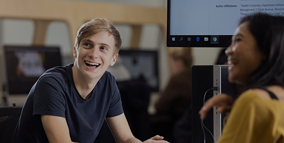 Two students sat in the library, laughing. One has their back to the camera and the other is looking off-camera.
