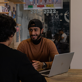 Two students sat opposite sides of a table with a laptop between them. The person facing the camera is smiling and wearing a cap