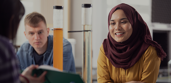 Close-up of a female student showing another female student something in a text book. They are sat in a lab.