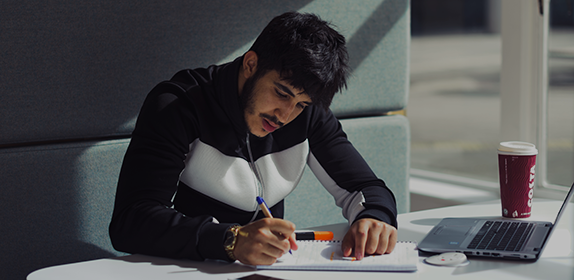 Close-up of an international student making notes in a notepad. He has his laptop open and it's a sunny day outside.