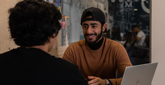 Tqo QA Pathway students sat talking. They have a laptop with them and it's open.