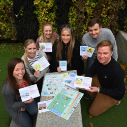 a group of people pictured sitting around a board game, holding up cards which are part of the game and smiling at the camera.