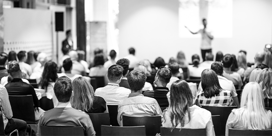 Image of a lecture theatre taken from the behind the audience with a speaker in the distance.