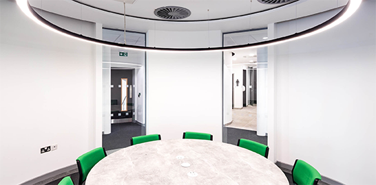 A white room with a circular table, green chairs and a large circular light fitting above. There are two doors leading out the room behind the table.