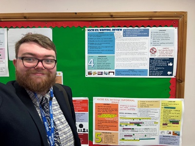 the image features Nick wearing a jacket and tie, standing in front of a notice board