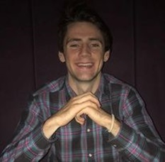 A man with black hair smiling. He is in a checked shirt with his elbows on a table with one hand on top of the other.