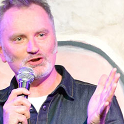 A man with grey hair and a beard on stage with a microphone. 
