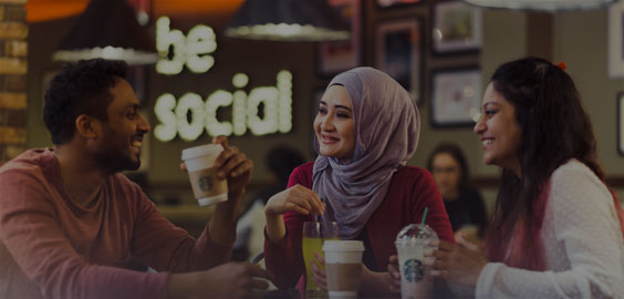 Image of a three students having drinks in Northumbria Students' Union with a Be Social light sign on the wall in the background
