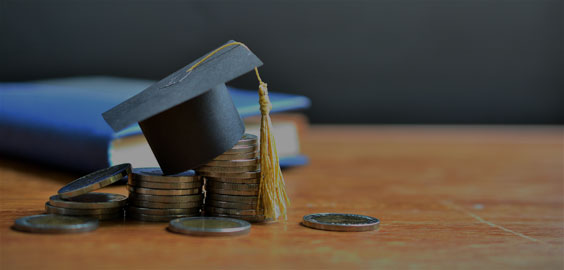 Image of a mini graduation cap resting on top of coins sitting on a wooden table with a blue book in the background